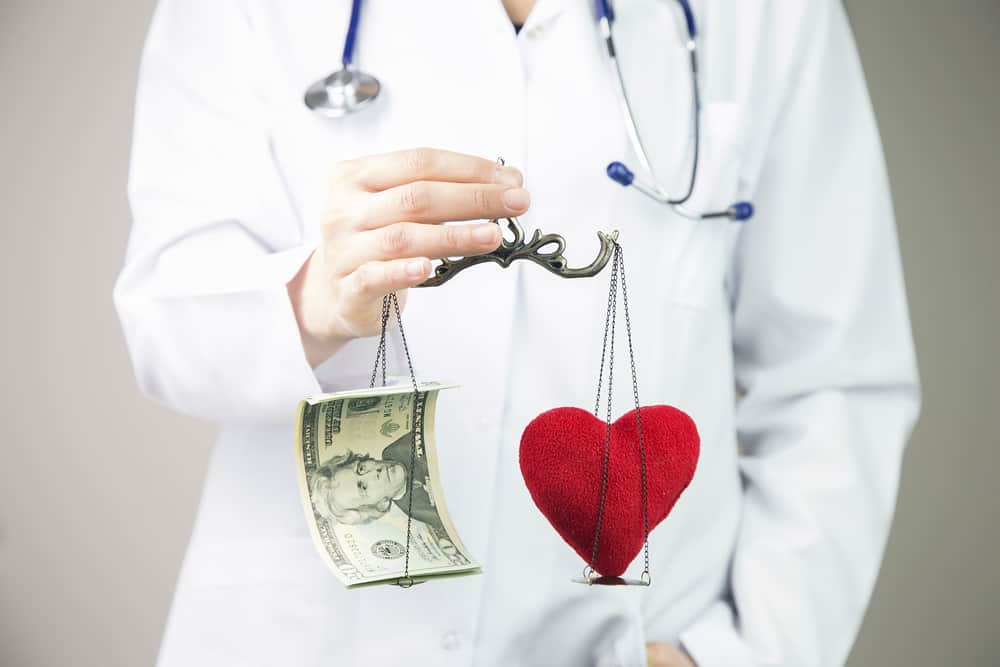 A doctor balances a $20 bill and a heart on a scale to show how understanding reference-based pricing works is crucial for saving money on healthcare coverage.