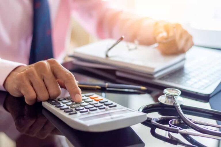 A man enters numbers on a calculator with a stethoscope lying on a desk to indicate healthcare payment integrity