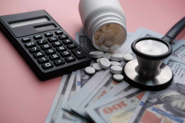 A calculator, stethoscope, and bottle of pharmaceuticals sit atop hundred dollar bills to show savings from managed care in healthcare