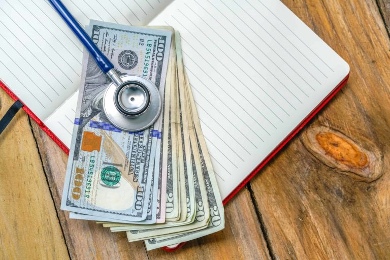 The value of cost containment in healthcare is illustrated by a stethoscope atop a pile of money and a blank journal on a wooden table
