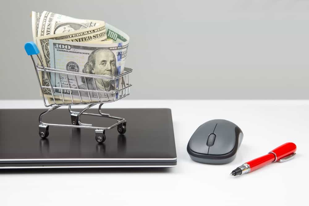 Strategies to reduce healthcare costs are represented by a roll of money in a miniature shopping cart atop a laptop.